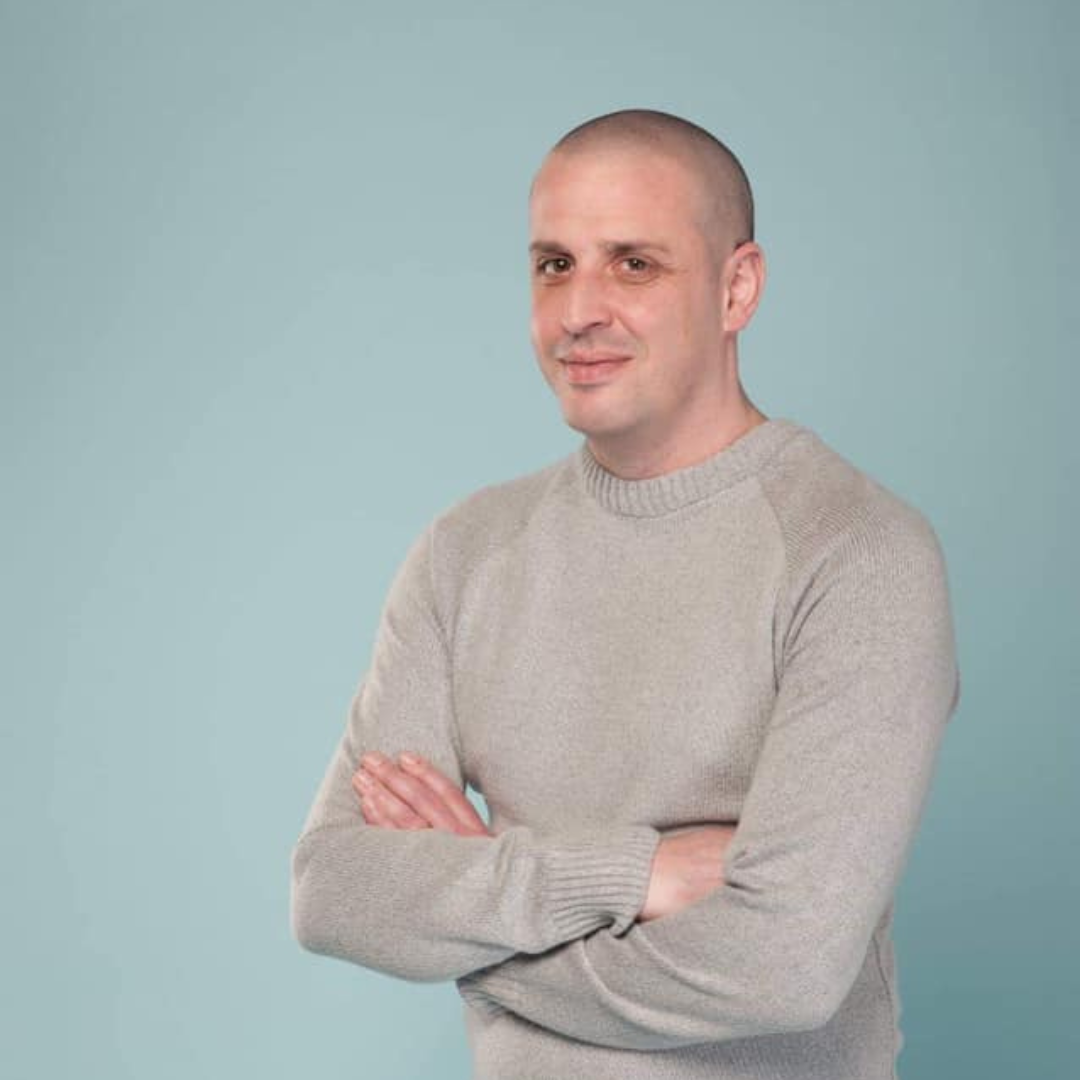 White male standing with his arms crossed in front of him. Wearing a beige sweater.